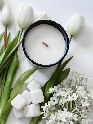 White candle in a black container surrounded by white tulips and flowers on a light background