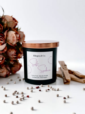 Matte black 8.5oz soy candle with copper lid and wooden wick styled in soft neutral decor, inspired by white musk and mimosa floral fragrance.